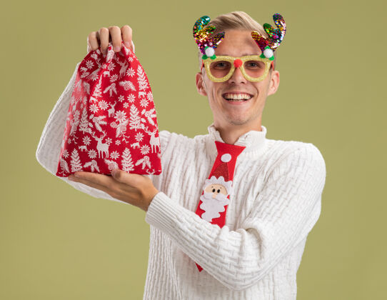 抱着快乐的年轻帅哥戴着圣诞新奇眼镜 打着圣诞老人的领带 手拿圣诞袋 看着橄榄绿背景上的相机圣诞老人绿色圣诞