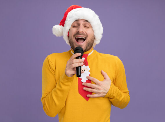 麦克风快乐的年轻白种人戴着圣诞帽打着领带拿着麦克风闭着眼睛唱歌手放在胸前紫色背景上隔离手年轻领带