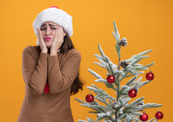 手戴着圣诞帽打着领带的年轻漂亮女孩站在圣诞树旁 双手放在橘色背景下孤立的脸颊上橘子帽子关心