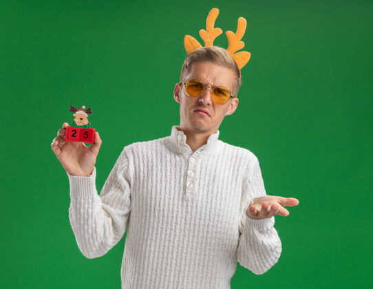 驯鹿皱眉的年轻帅哥戴着驯鹿鹿角头带戴着眼镜拿着圣诞树玩具和日期看着相机显示空手孤立在绿色背景上手日期眼镜