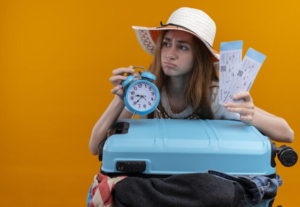橙色焦虑的年轻旅客女孩戴着帽子拿着飞机票和闹钟带着手提箱在孤立的橙色空间与复印空间女孩焦虑空间