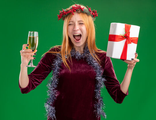 绿色兴奋的年轻漂亮女孩身穿红色连衣裙 脖子上戴着花环和花环 手里拿着一杯香槟 绿色背景上有一个孤立的礼盒圣诞快乐杯子花环