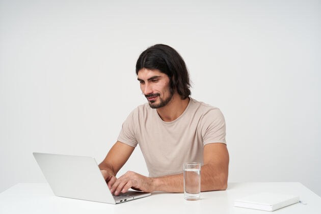欧洲人英俊 积极的商人的肖像 留着黑色的长发和胡须办公室概念专心工作在笔记本电脑上坐在白色的办公桌上工作场所 隔着白色的墙壁成人情绪工作