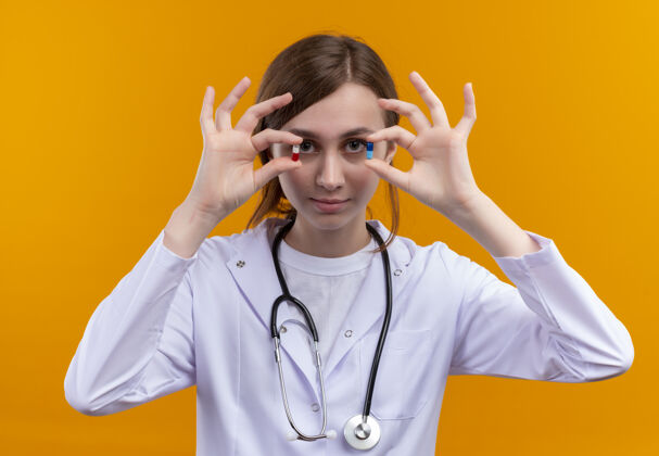 穿着自信的年轻女医生穿着医用长袍和听诊器在孤立的橙色空间展示医用胶囊胶囊展示听诊器