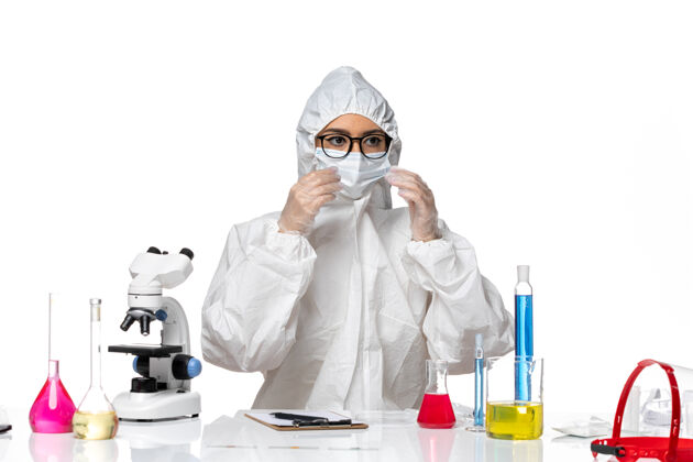 视图正面图：穿着特殊防护服的女化学家坐在白色办公桌上 手里拿着溶液化学大流行性冠状病毒外套女化学家药品