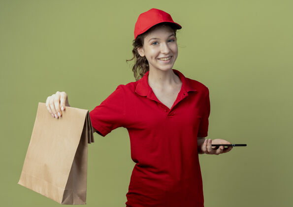 送货微笑着的年轻漂亮的送货女孩 穿着红色制服 戴着帽子 在橄榄绿的背景下 伸出一个纸包制服年轻女孩