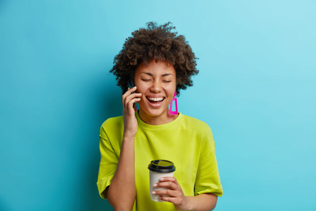 女性积极的非裔美国妇女通过手机交谈有愉快的交谈拿着外卖咖啡心情很好美国人复制女孩