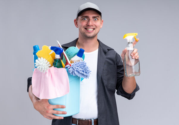 桶面带微笑的年轻帅哥 穿着t恤 戴着帽子 手里拿着一桶清洁工具和喷雾瓶 隔离在白色的墙上喷雾清洁家伙