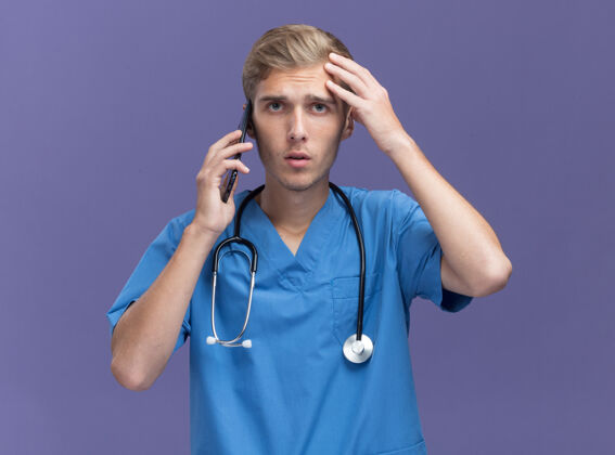 年轻穿着医生制服 手持听诊器的年轻男医生正在讲电话 手放在隔离在蓝色墙上的前额上说话不愉快手