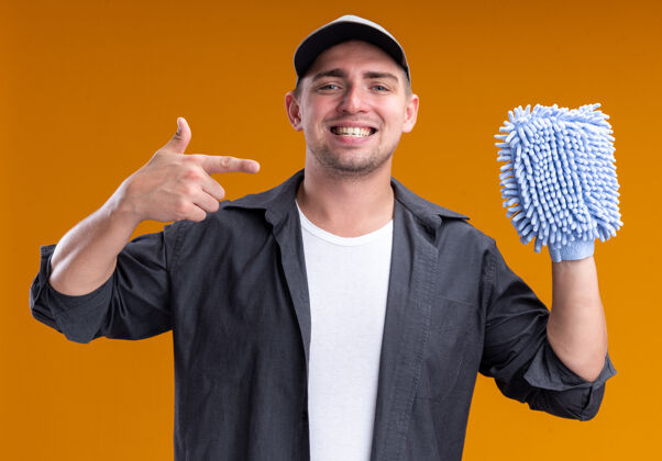 破布微笑着的年轻帅气的清洁工穿着t恤 戴着帽子 指着隔离在橙色墙上的抹布穿清洁年轻