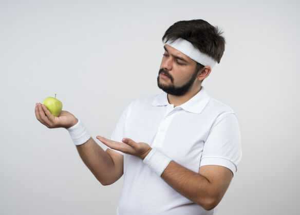 穿戴着头带和腕带 手拿苹果指着隔离在白墙上的一个未被释放的年轻人腕带点不愉快