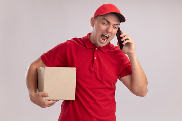 年轻愤怒的年轻送货员穿着制服 戴着帽子 拿着盒子 隔着白墙讲电话送货制服拿着