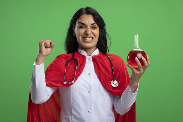 女孩快乐的年轻超级英雄女孩穿着医用长袍 听诊器拿着装满红色液体的化学玻璃瓶 在绿色的墙上显示“是”的手势液体手势医疗