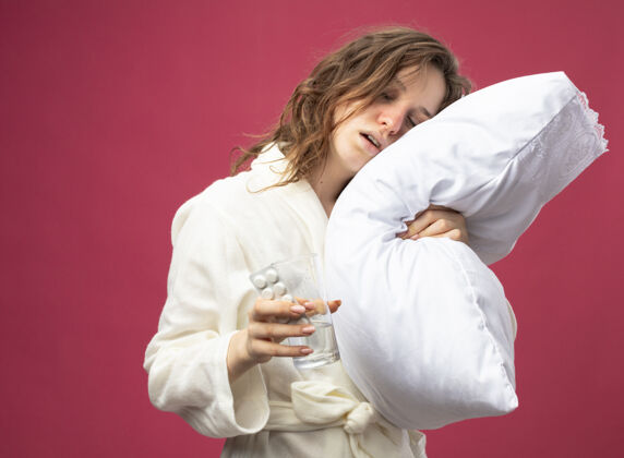 长袍虚弱的年轻病女孩穿着白色长袍抱着枕头拿着一杯水虚弱水女人