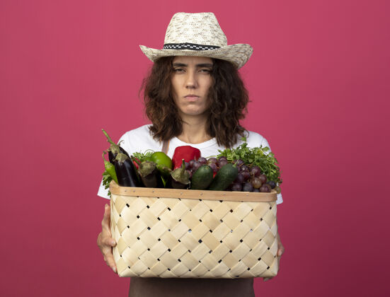 制服皱眉的年轻女园丁穿着制服戴着园艺帽拿着菜篮子人花园穿