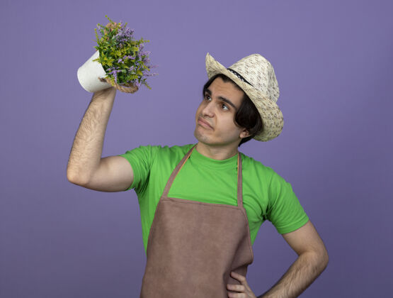 花盆穿着制服的年轻男园丁戴着园艺帽抬起头 看着花盆里的花 手放在屁股上 孤立地站在紫色上制服年轻花