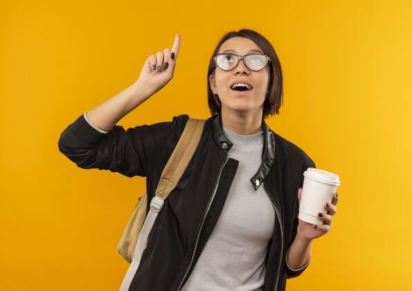 拿着印象深刻的年轻女学生戴着眼镜 背着袋子 拿着塑料咖啡杯 指着橘子孤立地仰望眼镜橘子印象