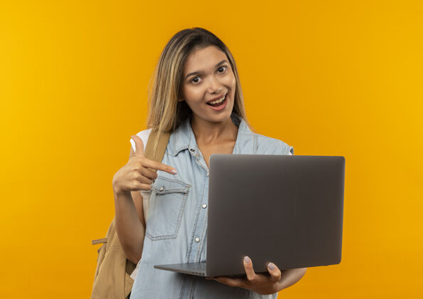 拿着快乐的年轻漂亮的女学生 背着书包 拿着笔记本电脑 指着橘黄色的笔记本电脑背橙色年轻