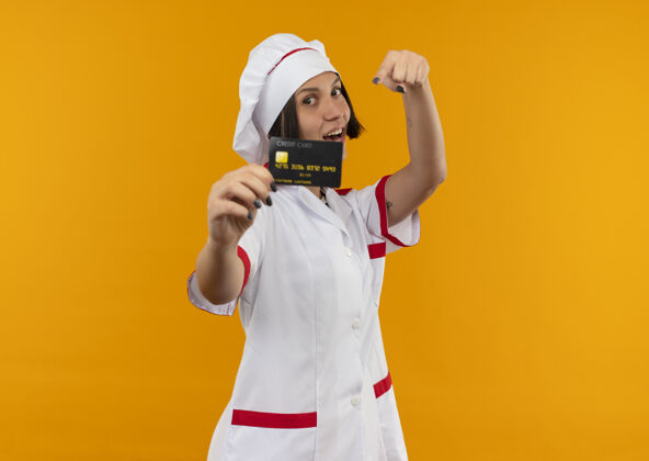 年轻身着厨师制服的快乐年轻女厨师伸出信用卡 指着橘子上孤立的信用卡走向烹饪指向