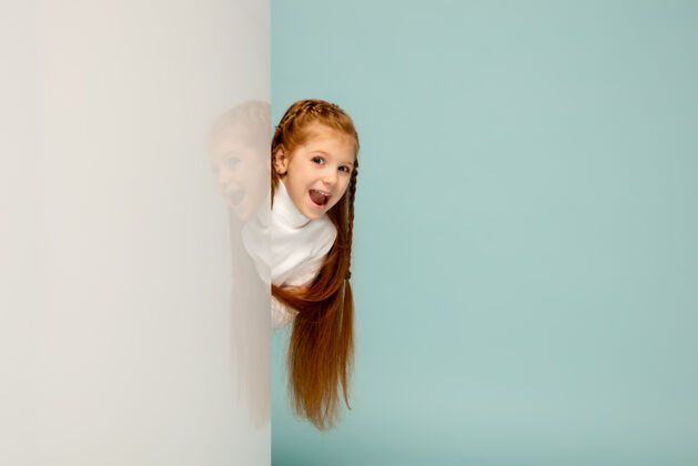 面部惊讶快乐的孩子 女孩在蓝色工作室背景上孤立看起来快乐 开朗广告空间童年 教育 情感 面部表情概念从墙后向外窥视微笑行动自由