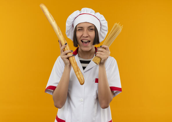 眨眼身着厨师制服的年轻女厨师 手里拿着面包棒和意大利通心粉 在橘子色的表面上相互隔离面包高兴意大利面