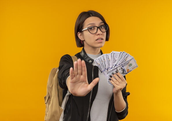 女孩未出租的年轻女学生戴着眼镜 背着包拿着钱 在橙色的背景上做着停止与复制空间隔离的手势不愉快穿抱