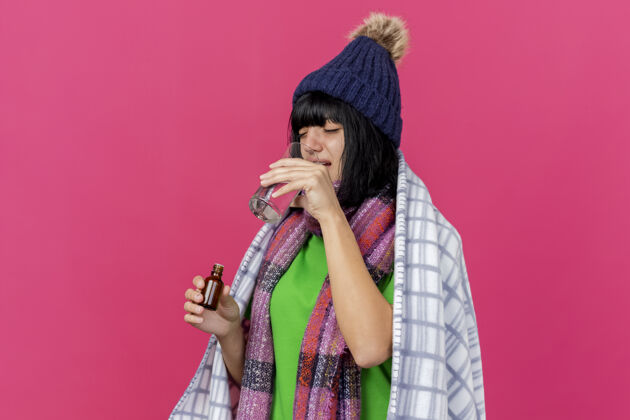 帽子年轻的生病妇女戴着冬天的帽子 围着方格布裹着的围巾 手里拿着装在玻璃里的药剂 喝着一杯水 隔离在粉红色的墙上 留着复印空间人喝酒脸