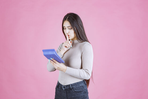 休闲穿着灰色衬衫的女孩手里拿着一个蓝色的计算器 一边工作一边打着沉默的手势计算器数学女性