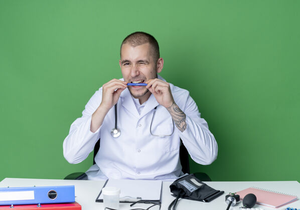 听诊器年轻的男医生穿着医用长袍和听诊器坐在办公桌旁 手里拿着工作工具 咬着笔 对着隔离在绿色背景上的摄像机眨眼眨眼年轻工作