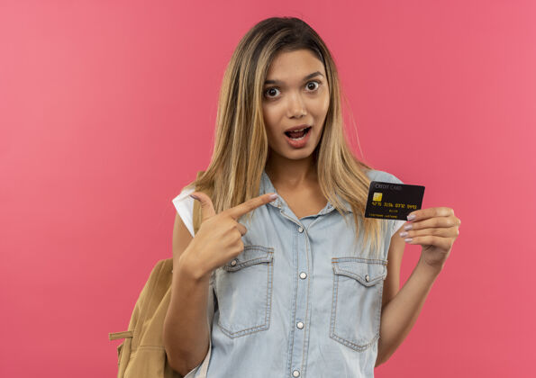 粉红印象深刻的年轻漂亮的学生女孩穿着背袋显示和指着信用卡孤立的粉红色学生年轻指向