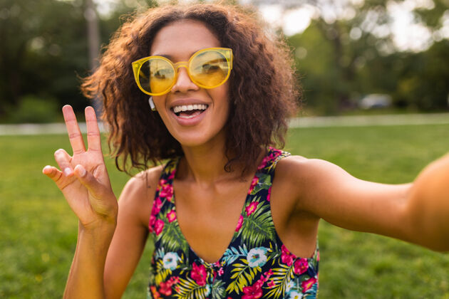 卷发年轻时尚积极的黑人女性 自拍 戴着无线耳机听音乐 在公园里玩得很开心 夏日时尚 五颜六色的时髦装扮混血儿无线自拍