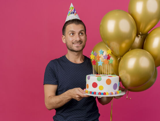 生日微笑英俊的白人男子戴着生日帽 拿着生日蛋糕和氦气球 看着在粉红色背景上与复制空间隔离的一面持有帽子蛋糕
