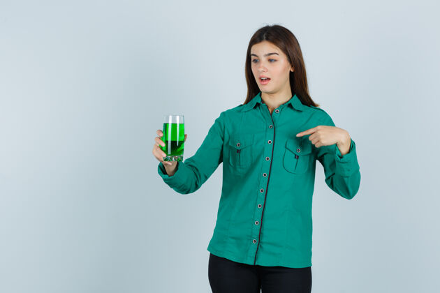 时尚年轻女孩穿着绿色上衣 黑色裤子 手里拿着一杯绿色液体 用食指指着它 一脸震惊 眼前一亮年轻女性卷曲