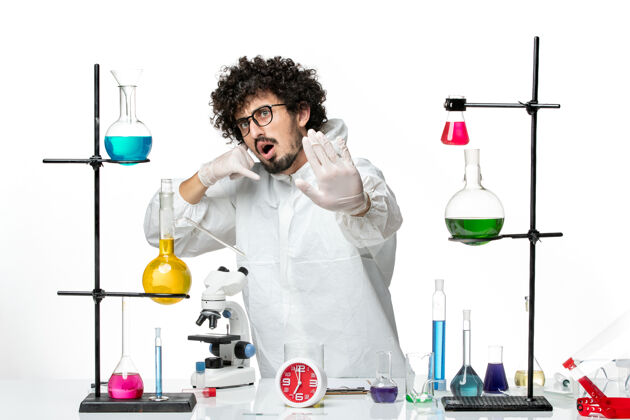 溶液正面图身着特殊套装的年轻男性科学家站在白色地板上 拿着解决方案围着桌子套装实验室化学