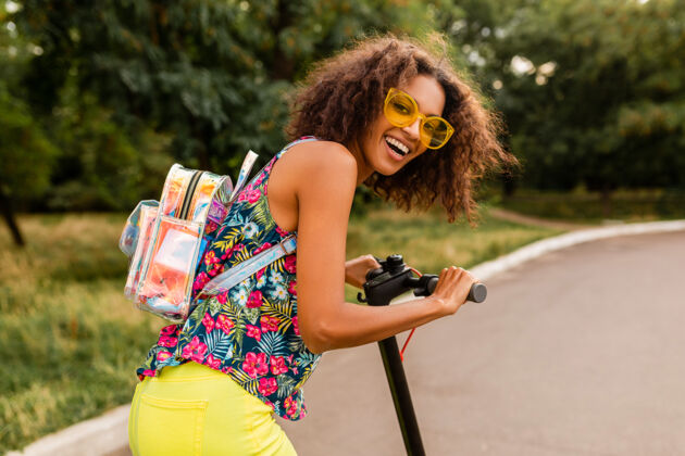 非洲裔美国人年轻时尚的黑人女子在公园里玩得很开心骑着电动脚踏车在夏天的时尚风格 五颜六色的时髦服装 穿着背包和黄色太阳镜车辆女孩环保