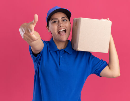 手势带着微笑的年轻送货员 穿着制服 肩上戴着帽子盒 在粉红色的墙上展示着你们的孤立姿态送货抱着盒子