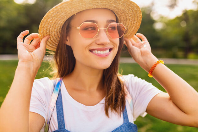 稻草戴着草帽 戴着粉色太阳镜 微笑着行走在公园里的年轻漂亮女人的肖像 夏日时尚的风格 五颜六色的时髦装扮年轻酷时尚