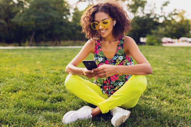 多彩年轻时尚微笑的黑人女子用智能手机听音乐无线耳机在公园里玩得开心 夏日时尚多彩的风格 坐在草地上 戴着黄色太阳镜黄色女士微笑