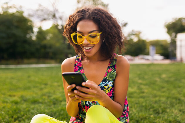 人年轻时尚微笑的黑人女子使用智能手机 通过无线耳机听音乐 在公园里玩得很开心黑色卷发年轻