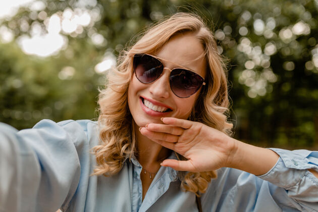 相机迷人的金发微笑的白牙女人穿着夏装在公园里散步在电话里自拍积极肖像眼镜