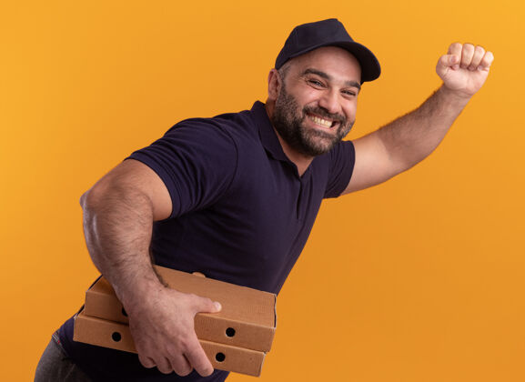 手势微笑着站在剖面图中年送货员制服和帽子举行比萨饼盒和显示运行的姿态孤立在黄色的墙壁上侧写盒子表演