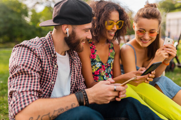 年轻快乐的年轻朋友们坐在公园里用智能手机微笑 男人和女人一起玩多样性时尚情绪