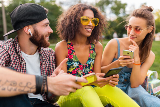朋友快乐的年轻朋友们坐在公园里用智能手机微笑 男人和女人一起玩太阳镜团体黑色