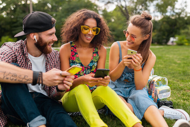 女人快乐的年轻朋友们坐在公园里用智能手机微笑 男人和女人一起玩时尚手机积极