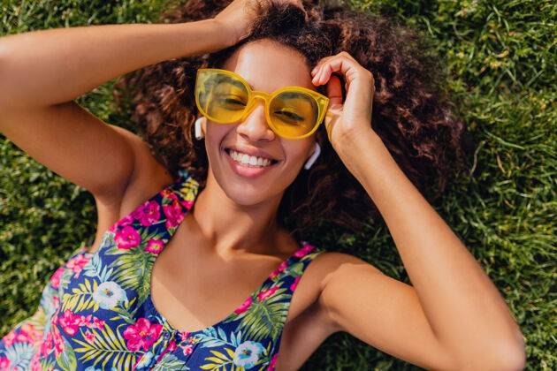 积极年轻时尚的黑人女子 戴着无线耳机听音乐 躺在公园的草地上玩得很开心 夏日的时尚风格 五颜六色的时髦装扮 俯瞰风景人物时尚人物