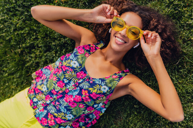 酷年轻时尚的黑人女子 戴着无线耳机听音乐 躺在公园的草地上玩得很开心 夏日的时尚风格 五颜六色的时髦装扮 俯瞰风景非洲裔美国人享受人物