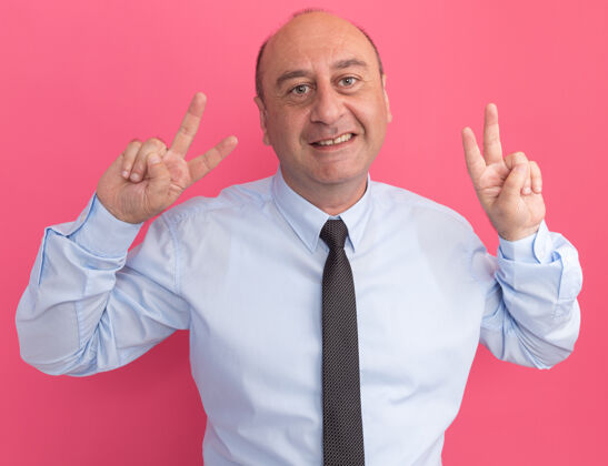 穿微笑的中年男子身穿白色t恤 打着领带 在粉红色的墙上显示出和平的姿态男人秀手势