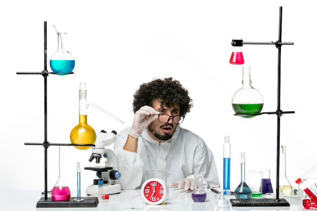 烧杯前视图穿着白色特殊套装的年轻男性科学家坐在那里 拿着解决方案 看着白墙上的东西 科学实验室里的男性化学医学特殊科学