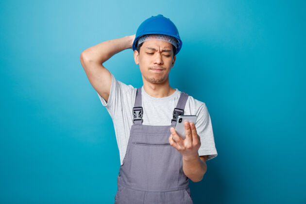 电话遗憾的是 年轻的建筑工人戴着安全帽 穿着制服 手放在脑后 拿着手机看蓝色建筑头盔