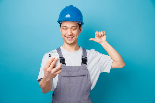 建筑面带微笑的年轻建筑工人戴着安全帽 穿着制服 拿着手机看着自己拿着安全电话